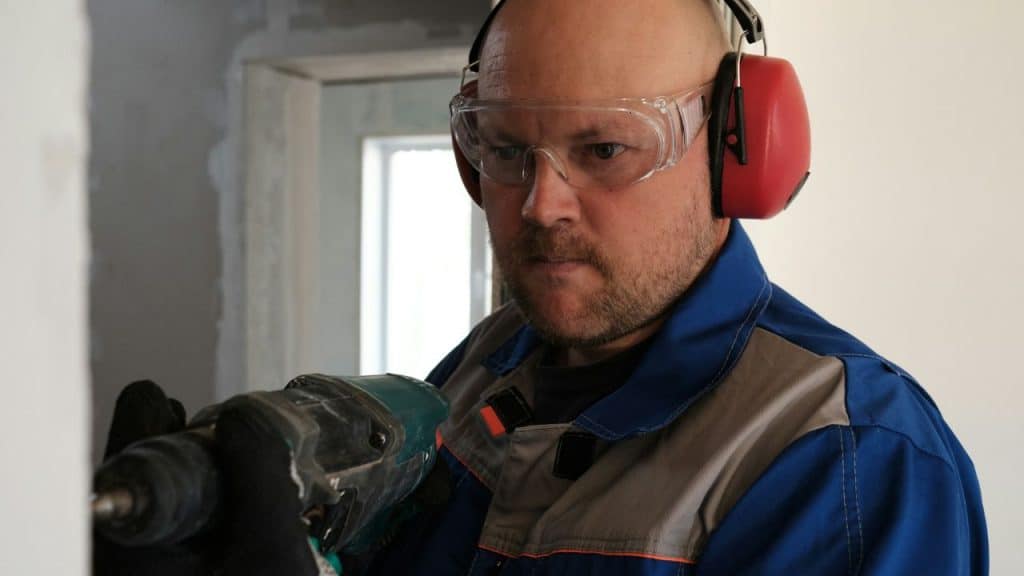A bald man in safety goggles and headphones drills into a wall.