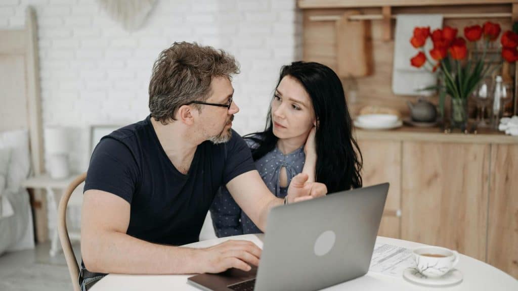 A couple sitting at a table talking while looking at a laptop.