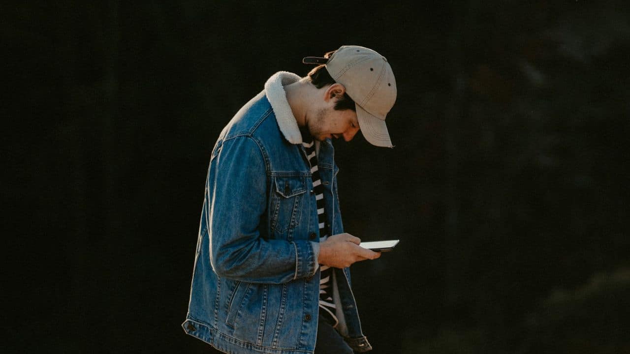 A man in a denim jacket and baseball cap looks down at a phone.