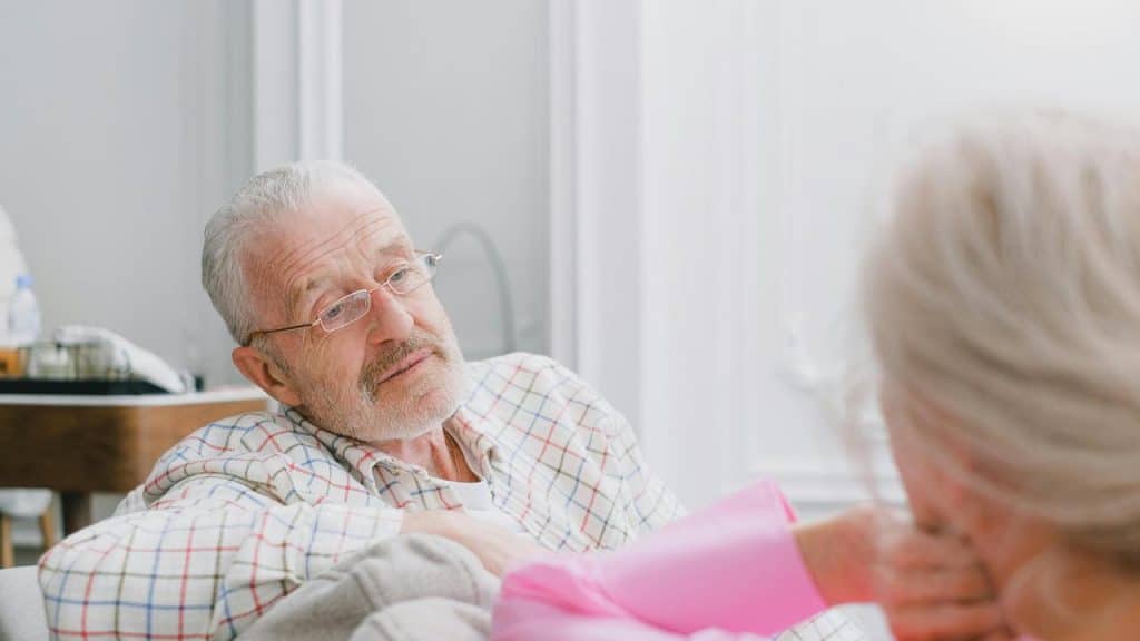 An elderly man wearing glasses talking with a woman in a bright room.