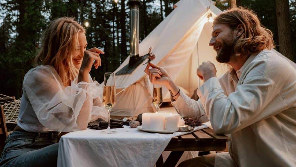 A couple enjoying a romantic outdoor dinner by a tent with candles and champagne.