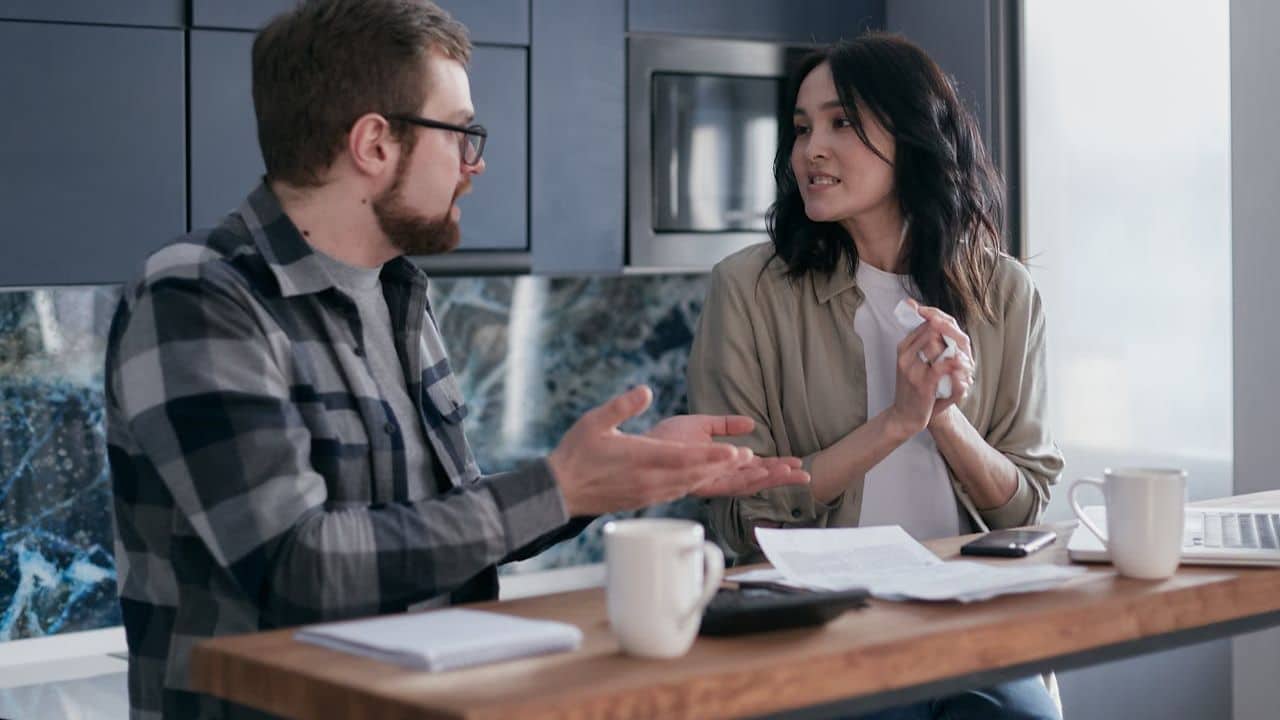 A man and woman sit at a kitchen counter, talking and gesturing during a discussion.