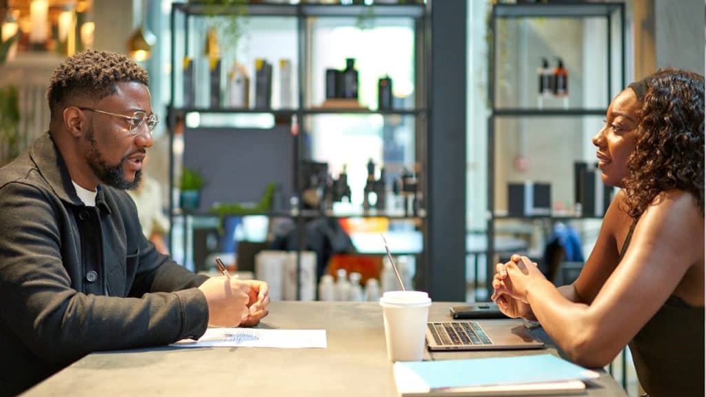 A man and a woman are sitting across a table from each other in an office or cafe, engaged in a professional conversation.