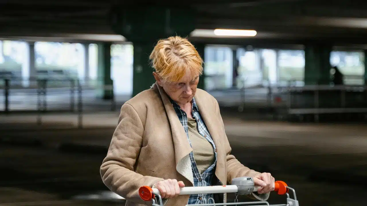 An older woman pushing a shopping cart in a dim parking garage.