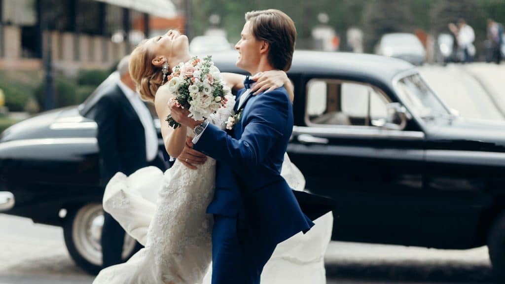 A bride in a white dress and a groom in a blue suit are embracing and dancing outside a vintage black car.