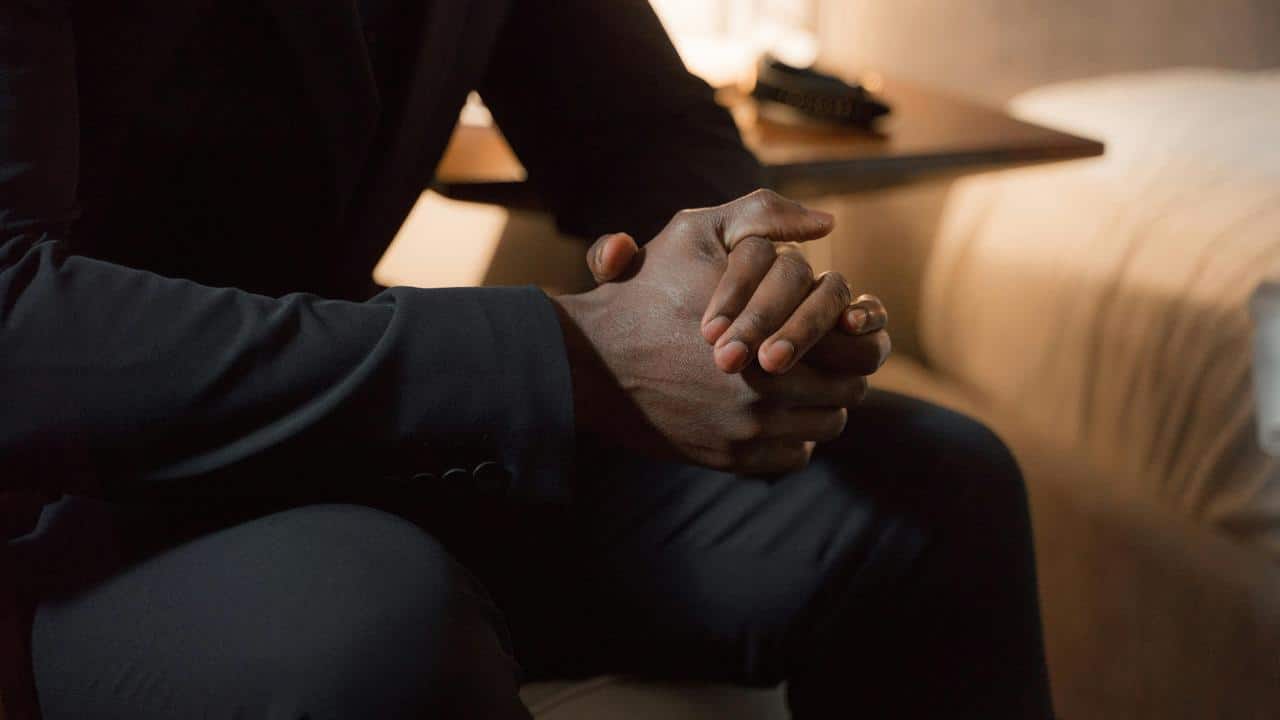A close-up of a man in a dark suit sitting with his hands clasped together.