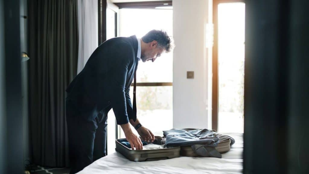 Middle-aged businessman packing a suitcase on a bed near a bright window.