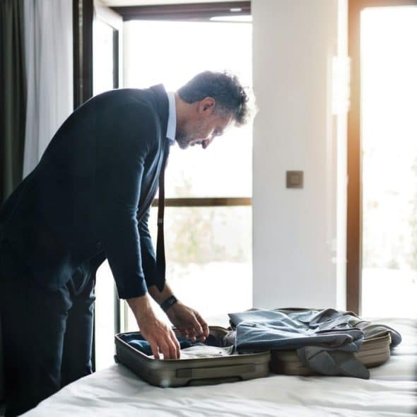 Middle-aged businessman packing a suitcase on a bed near a bright window.