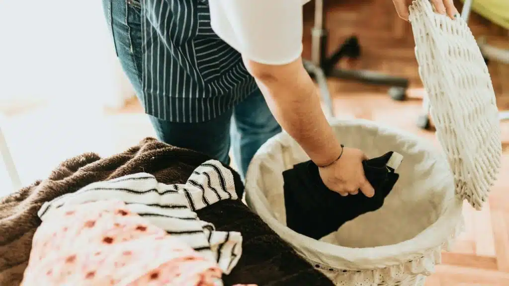 Person putting dark laundry into a white wicker basket with clothes nearby.
