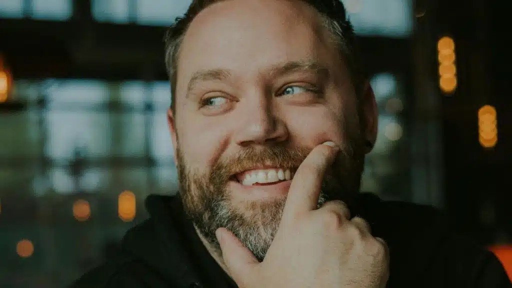 A bearded man with blue eyes is smiling and touching his chin.