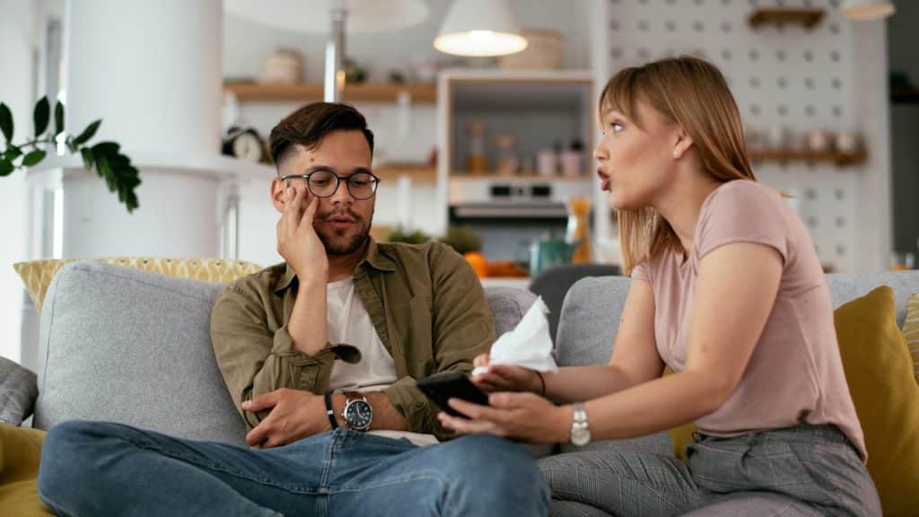 A woman is speaking emphatically to a man who looks distressed, as they sit on a couch in a living room.