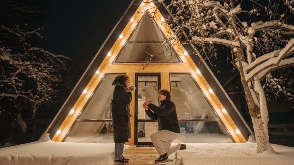 A man is proposing to a woman on his knee in the snow outside a brightly lit, modern A-frame house at night.