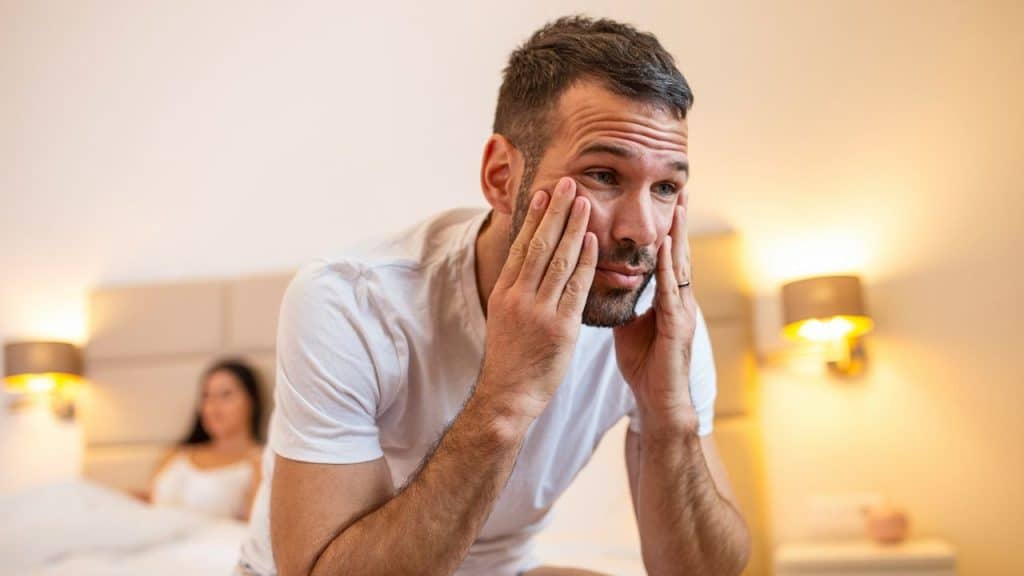 Distressed man sitting on a bed with his hands on his face, woman blurred in background.