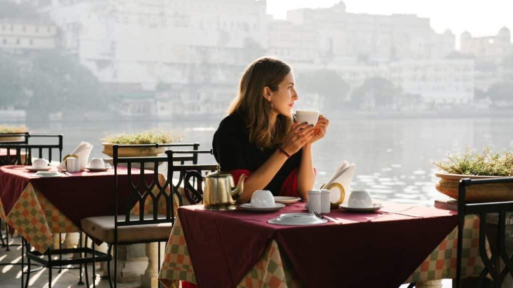 Woman holding a cup at an outdoor table overlooking a sunny waterfront cityscape.