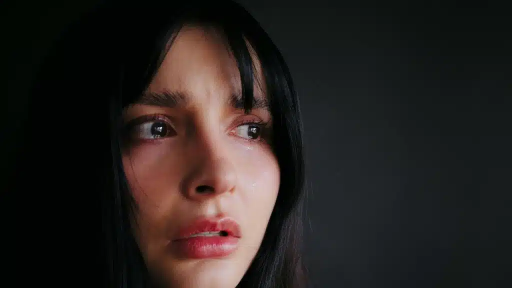 A woman with long dark hair looks distressed and tearful against a dark background.