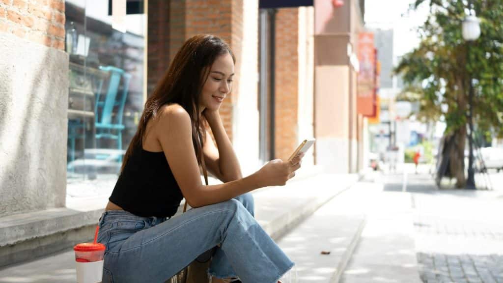 A woman sitting outdoors and smiling while looking at her phone.