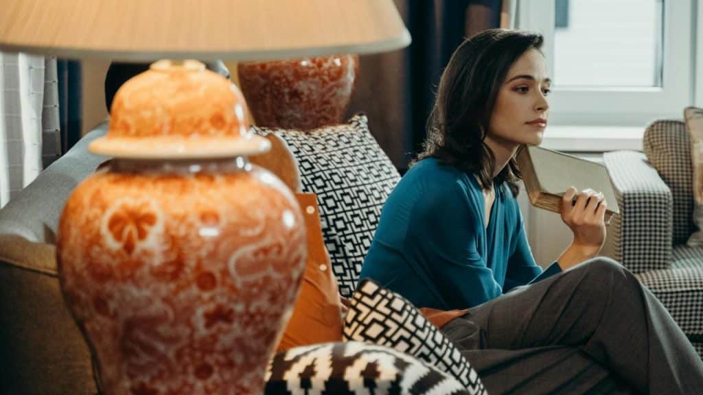 A woman in a blue top sits on a sofa holding a book and looking thoughtful.