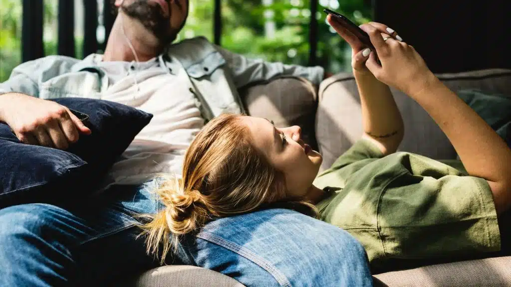 A woman lying on a couch with her head on a man’s lap, looking at her phone.