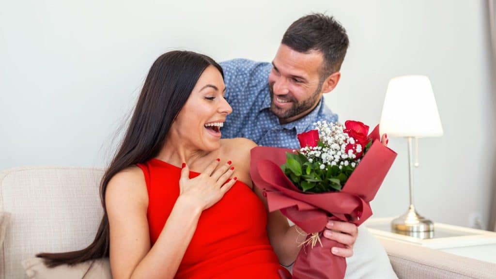 A smiling man presents an excited woman with a bouquet of red roses on a couch.