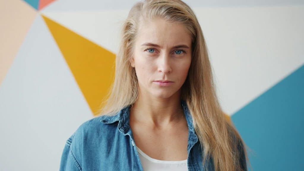 Blonde woman in denim shirt with neutral expression in front of geometric background.