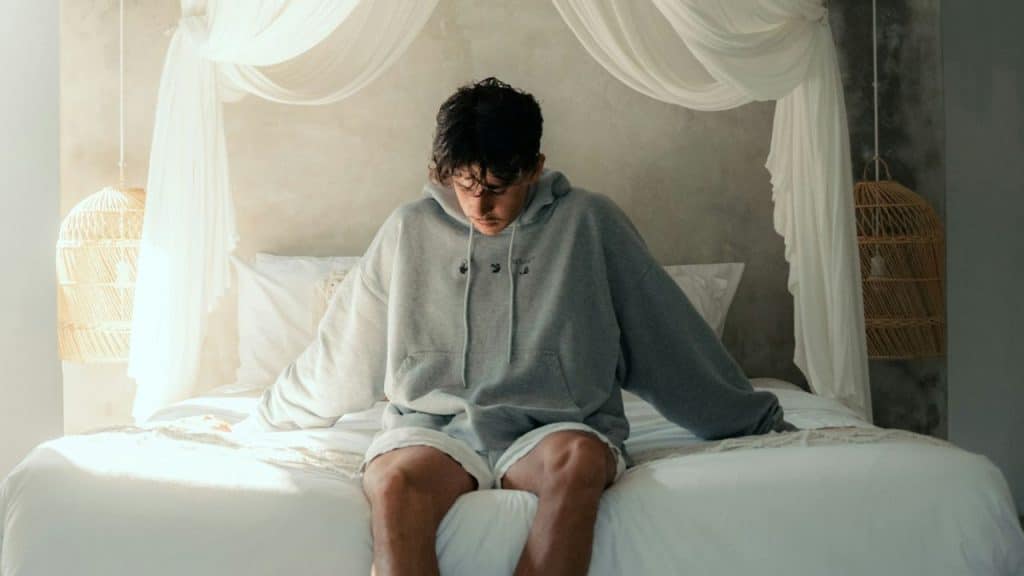 Young man in oversized gray hoodie sitting alone on a white bed with a canopy.