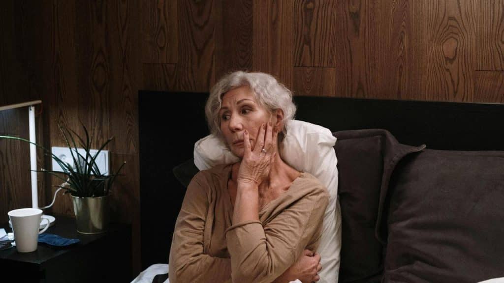 An elderly woman sitting on a bed with her hand on her face.