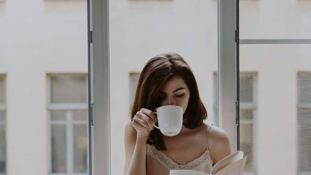 A woman sitting by a window, drinking from a cup while reading a book.
