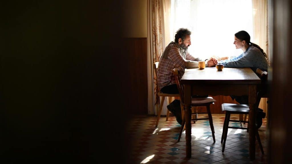 A man and a woman sit at a wooden table holding hands in a sunlit room.