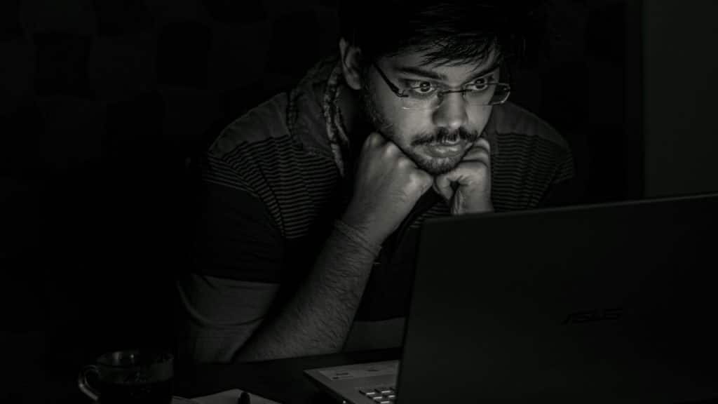A man with glasses rests his chin on his hands while staring intently at a laptop screen in the dark.