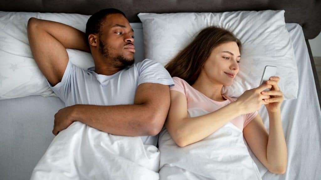 An overhead view of a man looking at a woman who is smiling while using her phone in bed.