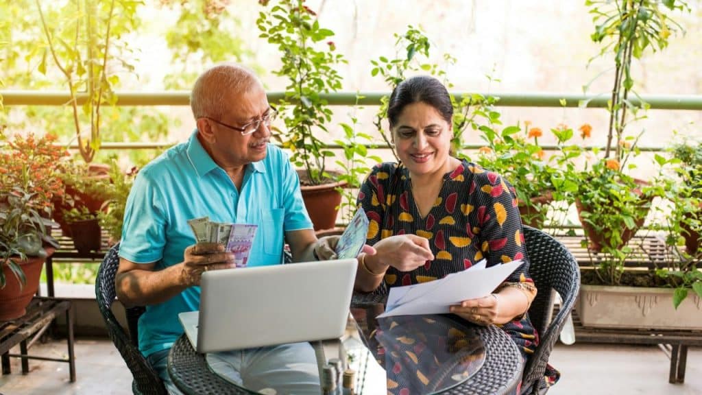 An older Indian couple on a balcony reviews a document, laptop, and Indian currency.