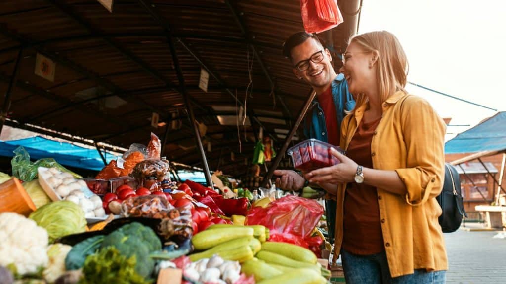 A smiling man and woman are shopping for produce at an outdoor farmers market stall filled with fresh vegetables.