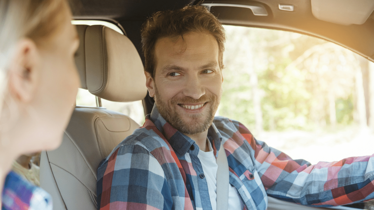 A smiling man in a plaid shirt is looking over his shoulder at a blonde woman while driving a car.