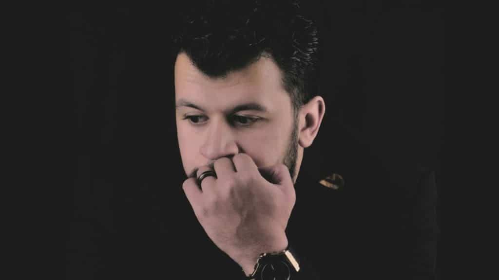A pensive man in a dark suit holds his fingers near his lips against a black background.