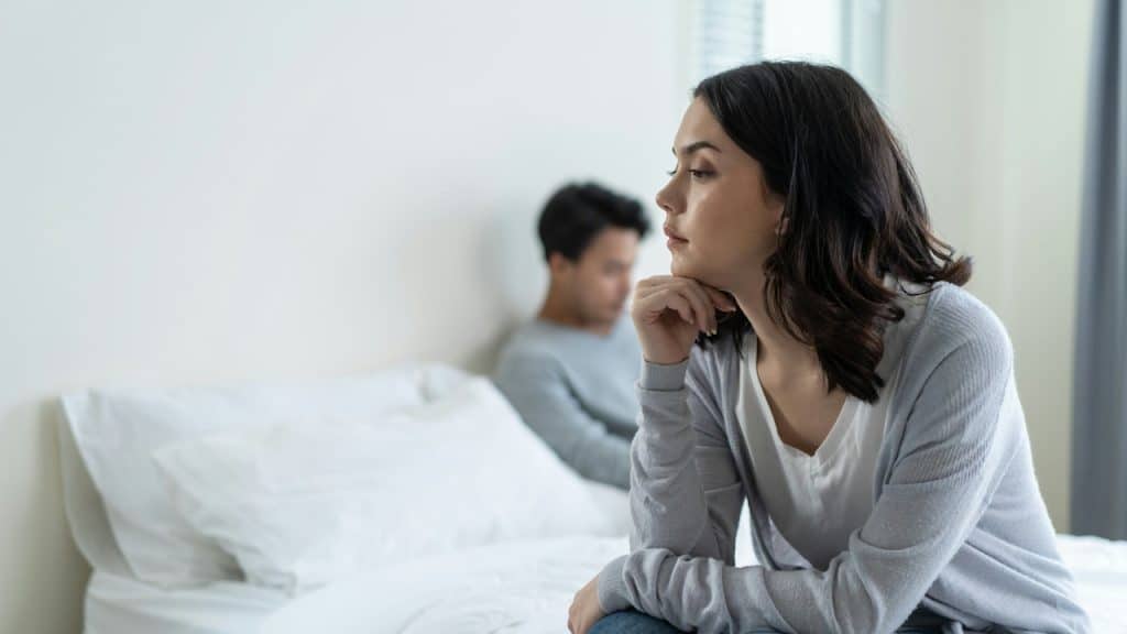A woman sits on the edge of a bed, looking away with a pensive or upset expression, while a man sits silently behind her.