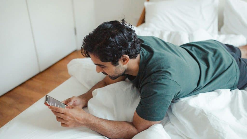 A man is lying on his stomach in bed, looking at his smartphone.