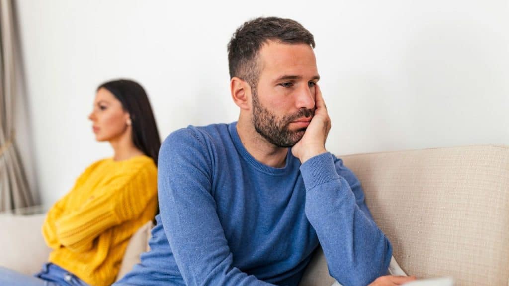 Distressed man sitting on a couch with a woman facing away from him in the background.