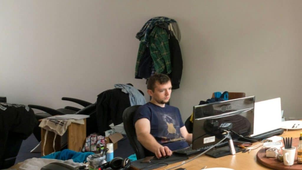 Man wearing a dark blue shirt intently working on a desktop computer in a messy room.