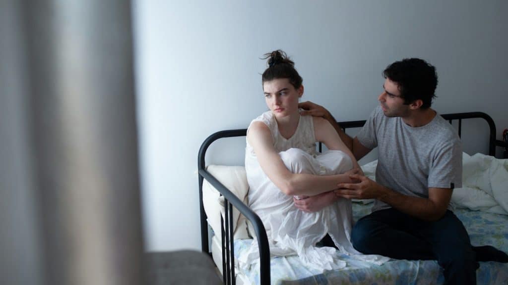 A man on a bed attempts to comfort a distressed woman who is sitting curled up and looking away from him.