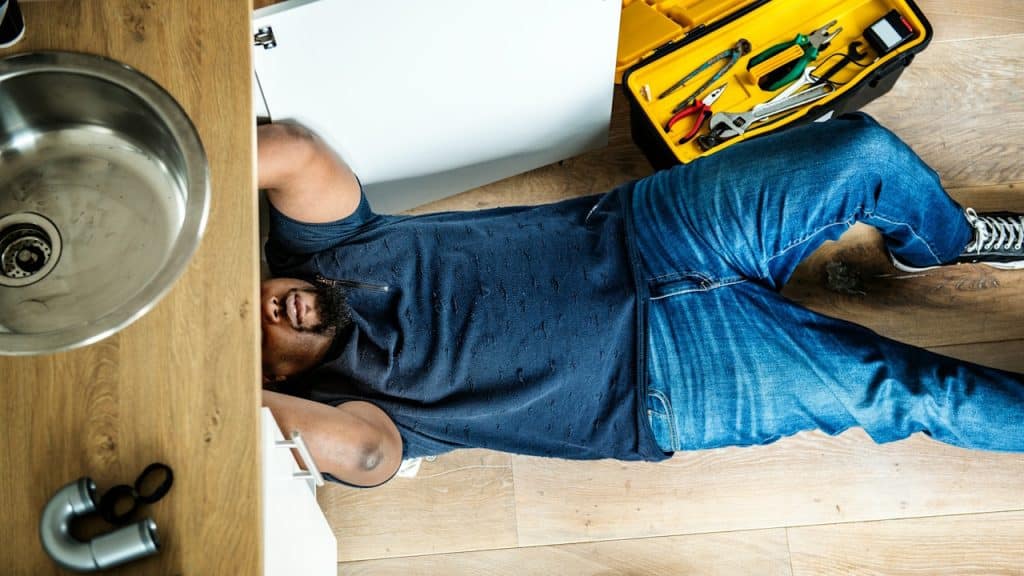 An overhead view of a man lying on a wooden floor, fixing a plumbing issue under a kitchen sink next to an open yellow toolbox.