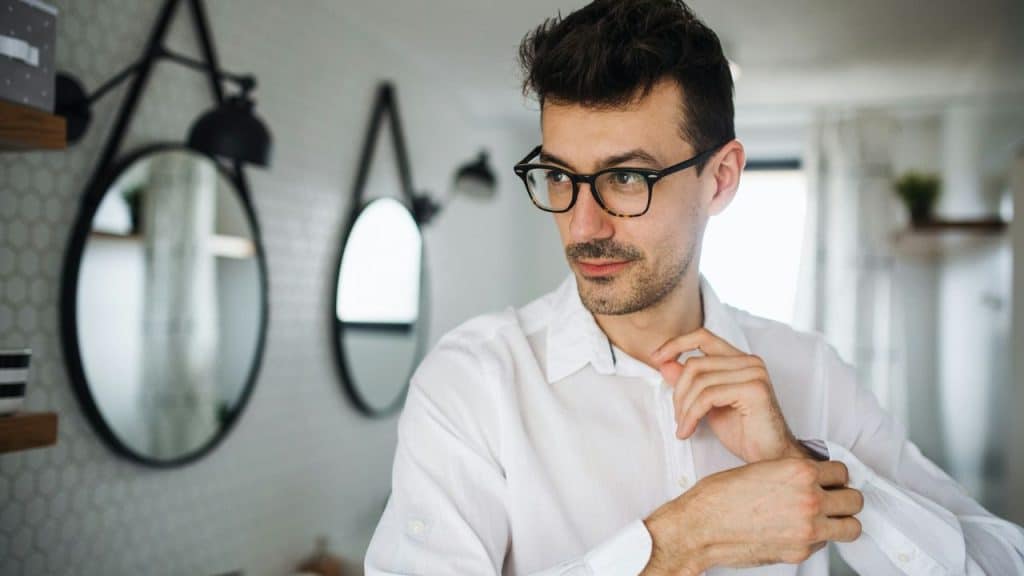 A man in glasses adjusts his white shirt in a room with round mirrors.