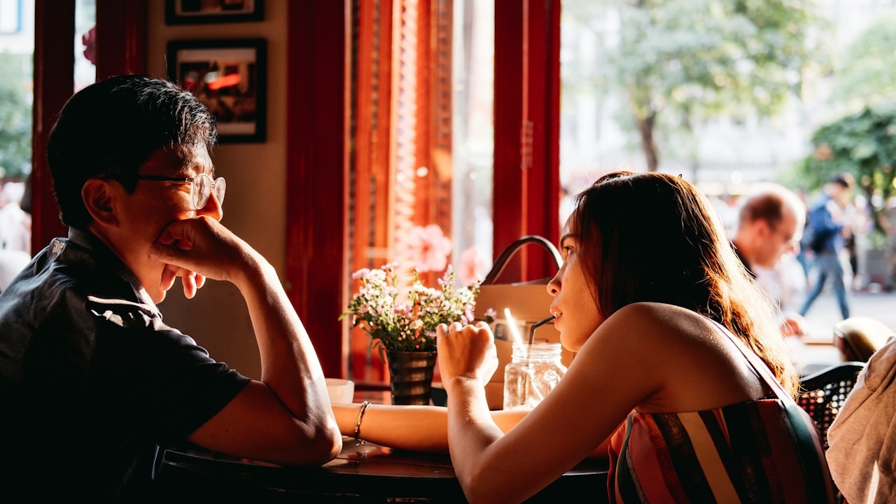 A man wearing glasses and a woman sit across a table from each other in a cafe with red trim, engaged in conversation.
