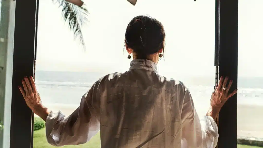 A woman standing by open doors looking out at the beach.