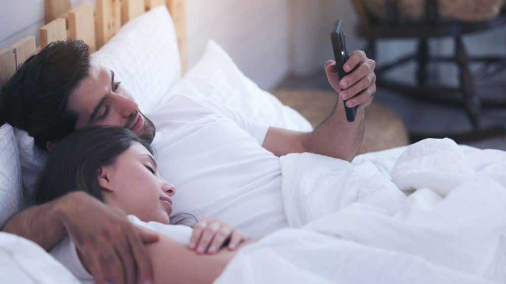 A bearded man is lying in bed, holding his phone while cuddling a woman who is sleeping next to him.