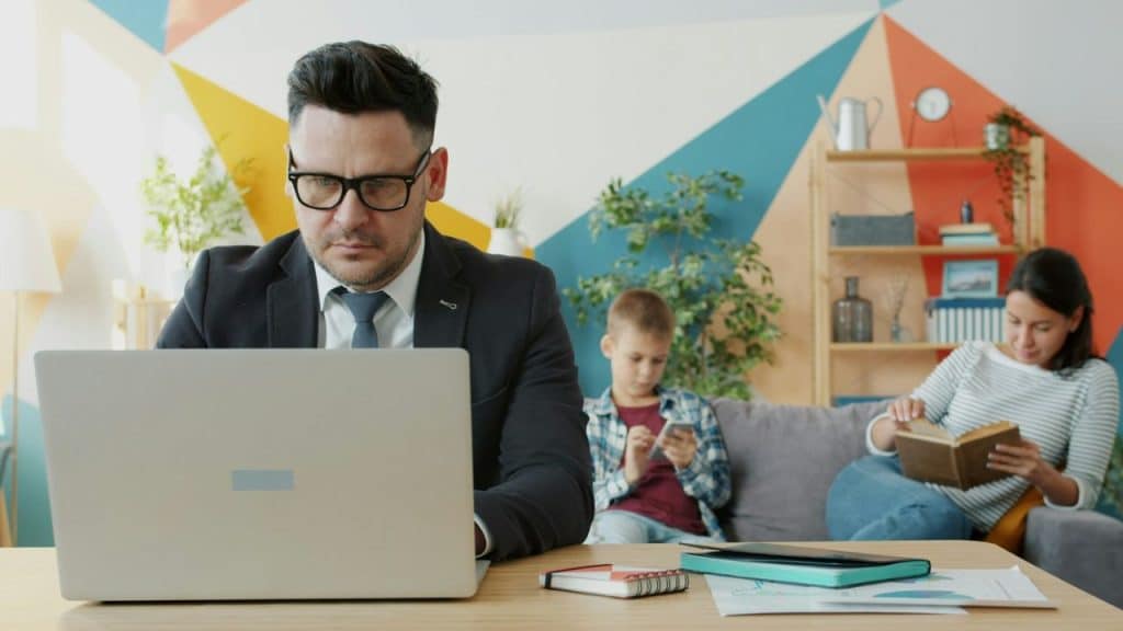 A father in a suit is engrossed in his laptop while his family sits behind him.