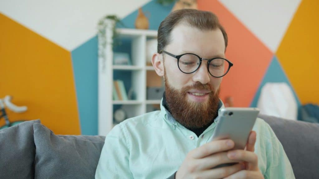 A smiling, bearded man with glasses is looking down at his smartphone in a brightly colored room.