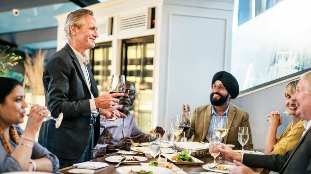 Mature man standing, holding a wine glass, talking to a group of friends at a dinner table.
