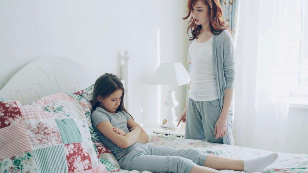 Woman pointing while talking to a young girl with arms crossed on a bed.