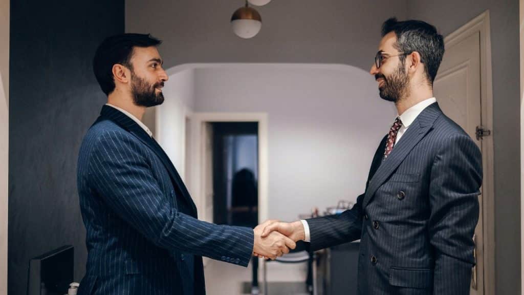 Two formally dressed men with beards shaking hands in an indoor hallway.