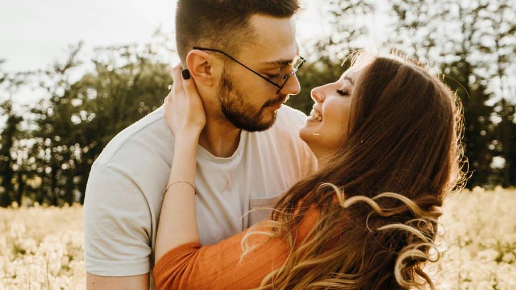 A couple leaning in close while smiling in a sunny field.